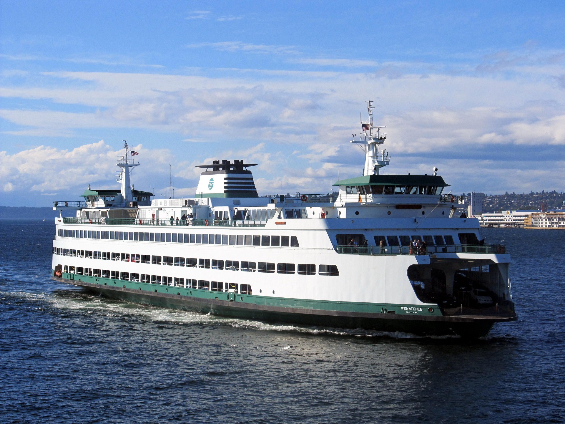 A Washington State Ferry cruising through Puget Sound under a clear blue sky, with the Seattle waterfront visible in the background.