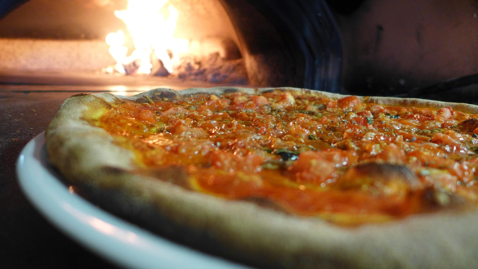 Wood-fired pizza with a golden, crispy crust and fresh tomato sauce, served on a white plate in front of a glowing brick oven.