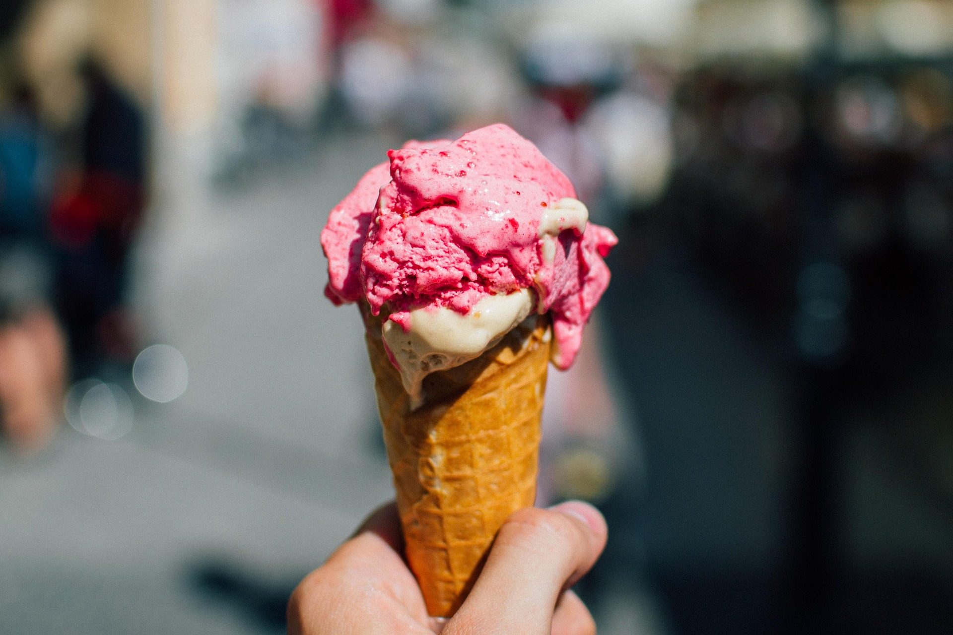 "A close-up of a melting double scoop ice cream cone with vibrant pink and creamy white flavors, held against a blurred city street background.